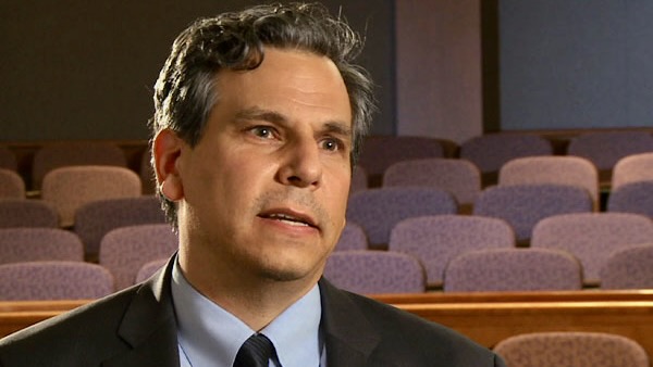 A man in a suit and tie, sitting in a theater, talking thoughtfully.