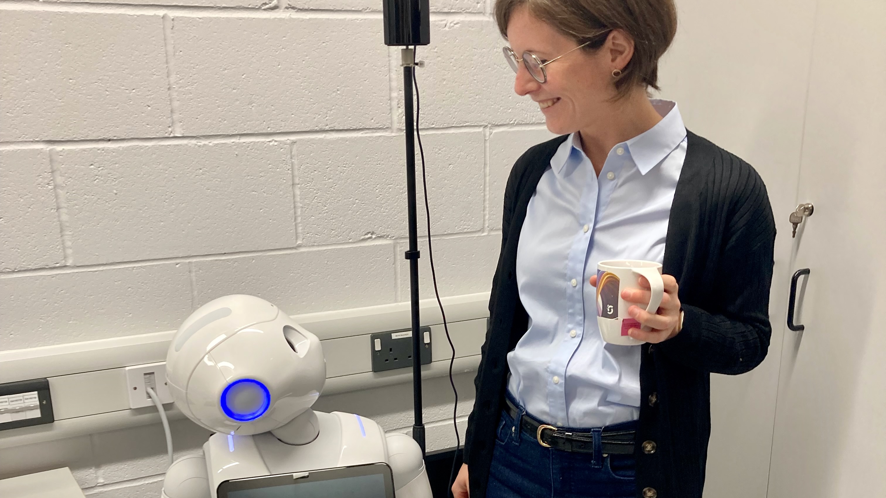 A young woman holding a coffee mug and smiling downwards at a robot.